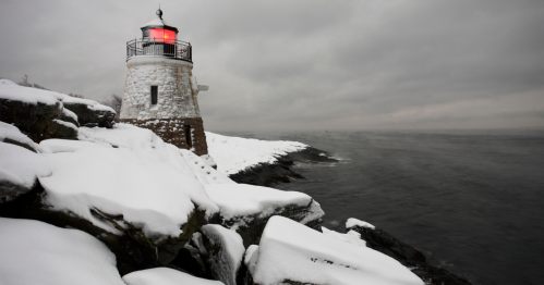 A snowy lighthouse stands on rocky shores under a cloudy sky, with a red light glowing at the top.