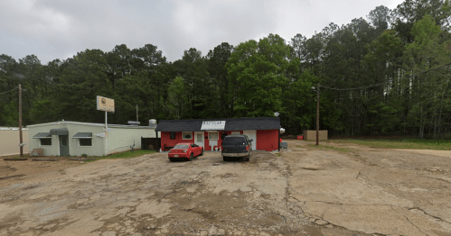 A small red building with a sign, surrounded by trees and parked cars on a gravel lot.