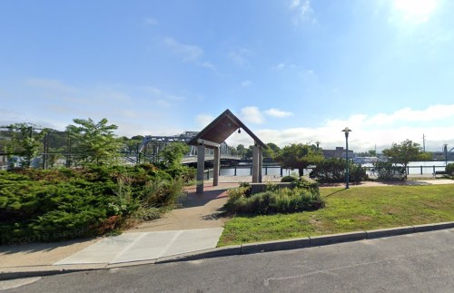 A park area with a gazebo, greenery, and a view of a bridge and water under a clear blue sky.
