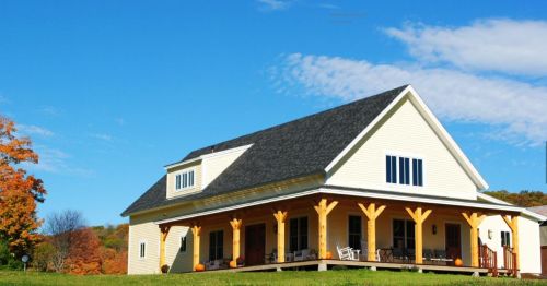 A large yellow house with a porch, surrounded by green grass and autumn trees under a clear blue sky.