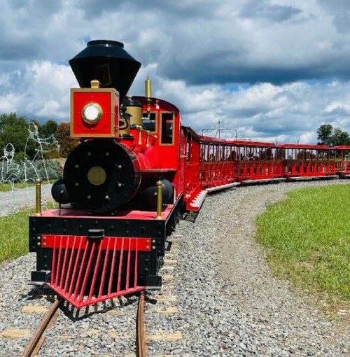 A bright red miniature train on a curved track, surrounded by green grass and a blue sky with clouds.