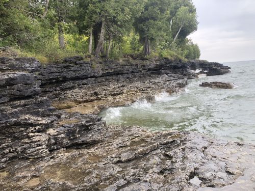Rocky shoreline with gentle waves lapping against the stones, surrounded by lush green trees and a cloudy sky.