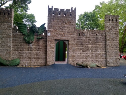A playground castle entrance with a dragon sculpture and a lizard figure on the ground, surrounded by greenery.