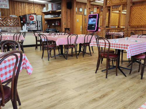 A cozy restaurant interior with wooden walls, checkered tablecloths, and empty tables. A soda machine is visible in the background.