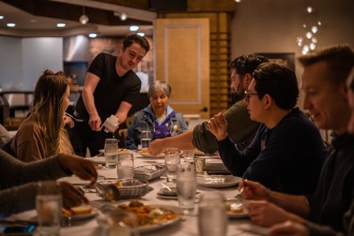 A waiter serves food to a group of people dining at a table in a cozy restaurant setting.