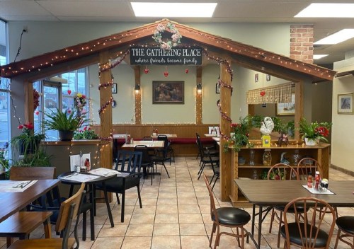 Cozy restaurant interior with wooden decor, plants, and a sign reading "The Gathering Place" adorned with lights.