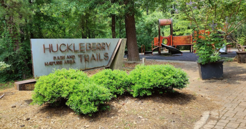 Sign for Huckleberry Park and Trails, surrounded by greenery, with a playground visible in the background.