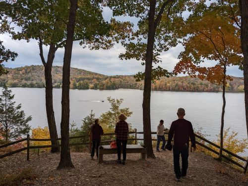 A serene lakeside view with autumn foliage, featuring people standing near a bench and a boat on the water.