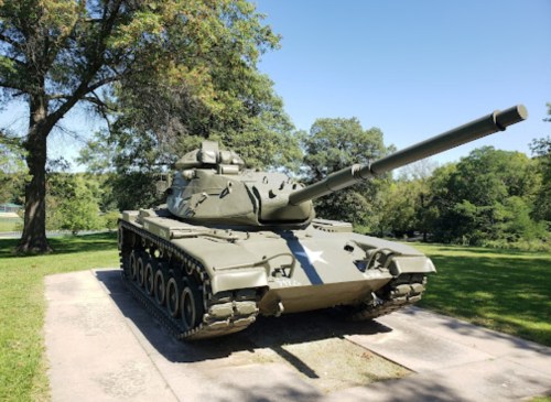 A military tank displayed on a concrete platform, surrounded by trees and grass under a clear blue sky.