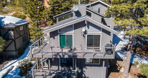 Aerial view of a modern gray cabin surrounded by trees and snow, featuring a large deck and multiple windows.