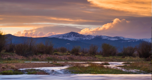 A serene landscape featuring a river, grassy banks, and mountains under a colorful sunset sky.