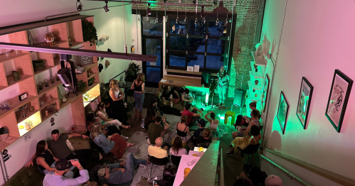 A lively indoor event with a crowd seated, green lighting, and a performer on stage in a cozy venue.
