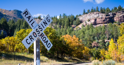 A railroad crossing sign stands in front of colorful autumn trees and rocky hills under a clear blue sky.