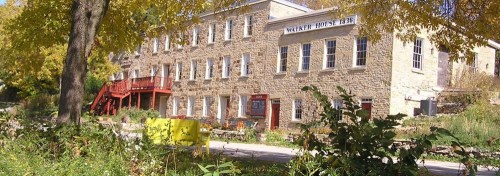 Historic stone building with red accents, surrounded by greenery and trees, featuring multiple windows and a welcoming entrance.