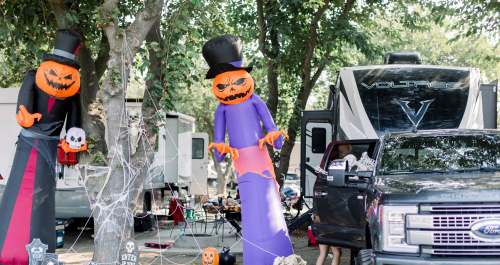 Two large Halloween decorations with pumpkin heads stand near a truck and RV, surrounded by festive decor and spider webs.