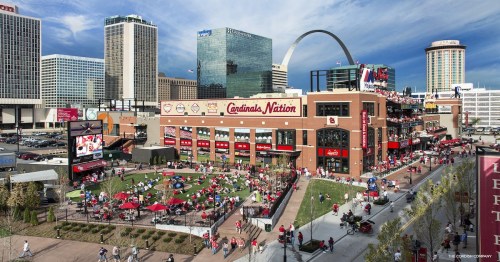 A vibrant outdoor area with fans in red, surrounded by buildings and the Gateway Arch in St. Louis.