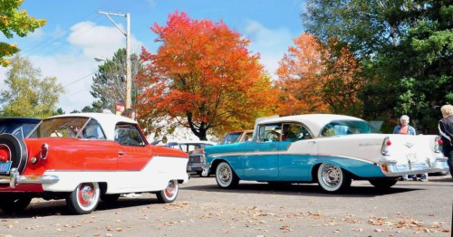Two classic cars, one red and white and the other turquoise and white, parked under vibrant autumn trees.