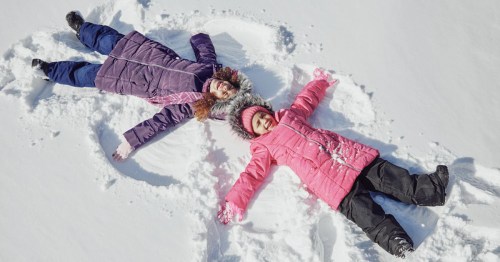 Two children in colorful winter coats making snow angels in a snowy landscape.