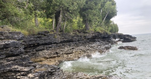 Rocky shoreline with gentle waves lapping against the stones, surrounded by lush green trees and a cloudy sky.