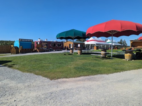 A food truck and picnic area with large umbrellas, surrounded by green grass and wooden structures under a clear blue sky.