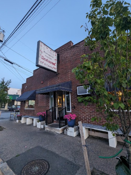 A brick restaurant exterior with a sign, potted flowers, and a blue awning, set in a quiet street at dusk.