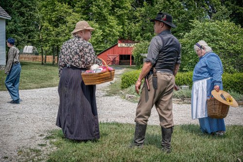A group of people in historical clothing, carrying baskets, conversing outdoors near a barn and greenery.