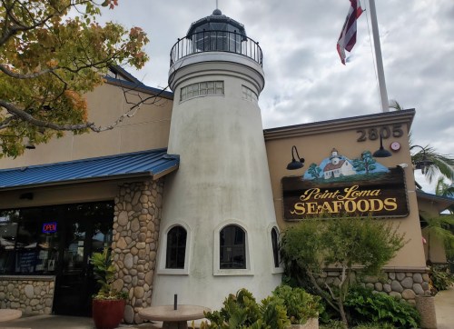 Exterior of Point Loma Seafoods featuring a lighthouse structure and a sign, with palm trees and a cloudy sky.