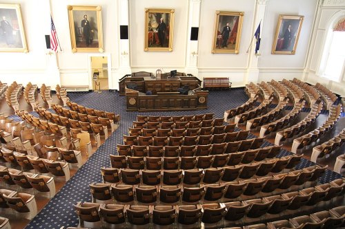 A spacious legislative chamber with rows of wooden seats, large portraits on the walls, and an American flag.