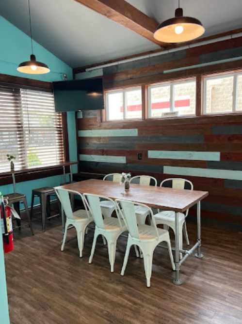 A cozy dining area with a wooden table, white chairs, and teal walls, featuring large windows and pendant lights.