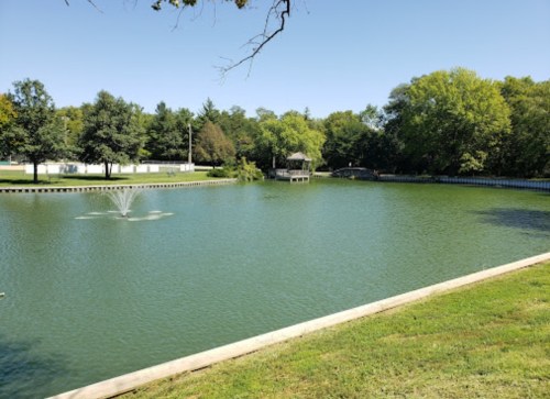 A serene park scene featuring a calm pond, fountain, and lush green trees under a clear blue sky.