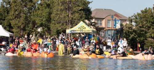 A lively scene of people in pumpkin boats on a lake, with spectators and a tent in the background.