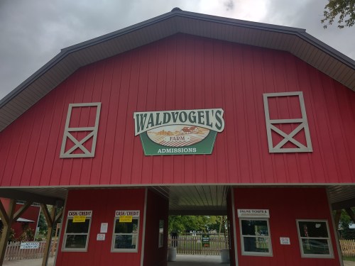 Red barn entrance with "Waldvogel's Farm Admissions" sign and ticket windows. Overcast sky in the background.
