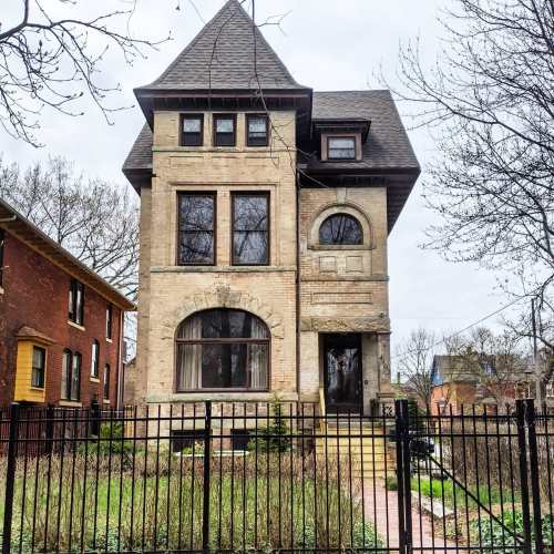 A tall, historic brick house with a pointed roof, surrounded by a black fence and trees in a residential area.