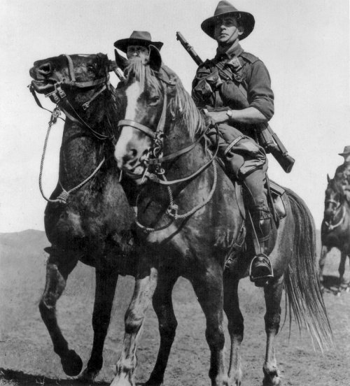 A soldier on horseback, armed with a rifle, rides alongside another horse and rider in a historical black-and-white photo.
