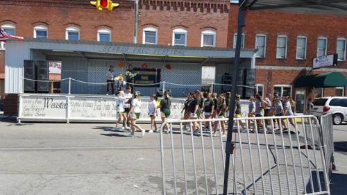 A group of people walking past a stage at a festival, with buildings and a barrier in the foreground.