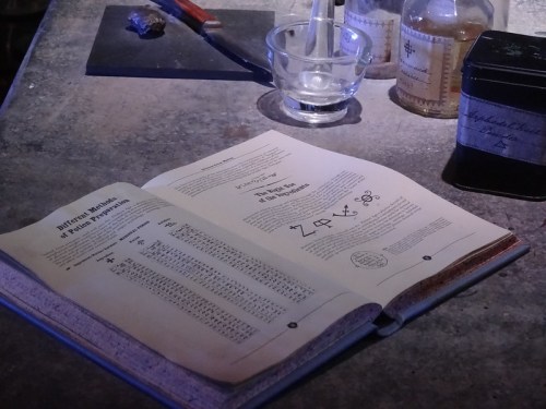 An open book on a table, displaying tables of symbols and text related to alchemy or potion-making.