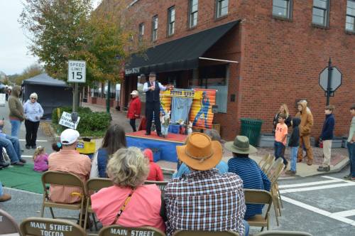 A crowd watches a performance on a small stage in a town square, with colorful decorations and a sign reading "Super Fun."