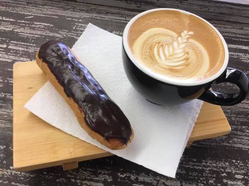 A chocolate-covered eclair next to a latte with a leaf design, served on a wooden tray with a napkin.