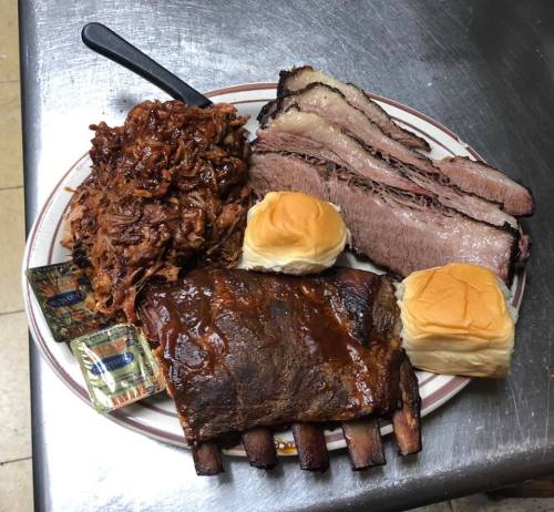 A plate of barbecue featuring ribs, brisket, pulled pork, and two dinner rolls with sauce packets on the side.