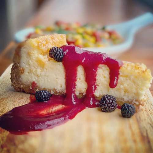 A slice of cheesecake topped with berry sauce and blackberries, served on a wooden board.
