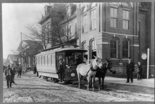 A historic black-and-white photo of a horse-drawn streetcar with passengers, set in a town with brick buildings.