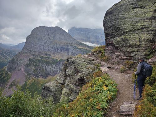A hiker stands on a rocky trail overlooking a mountainous landscape with lush greenery and cloudy skies.