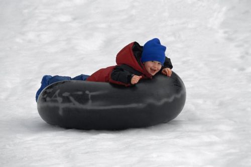 A joyful child in a blue hat and red jacket sleds down a snowy hill on a black inner tube.