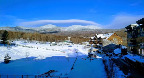 A snowy landscape with mountains in the background, blue sky, and a few buildings in a winter resort setting.