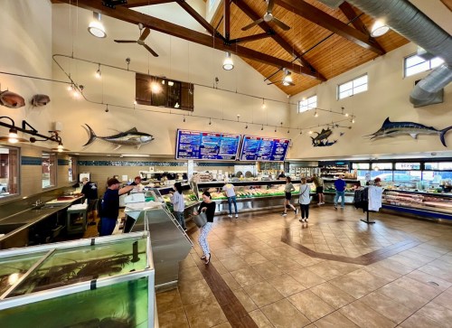 A bustling seafood market with customers browsing displays and a menu board, featuring a high ceiling and wooden beams.