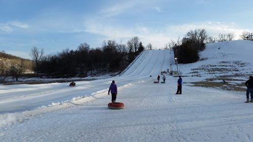 People tubing down a snowy hill on a winter day, with trees in the background and a clear blue sky.