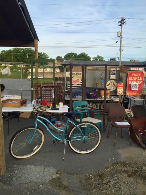 A cluttered outdoor space with a bicycle, tables, chairs, and various items for sale, surrounded by a fence.