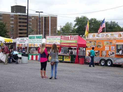 Two people stand in front of food stalls at an outdoor fair, with colorful tents and banners in the background.