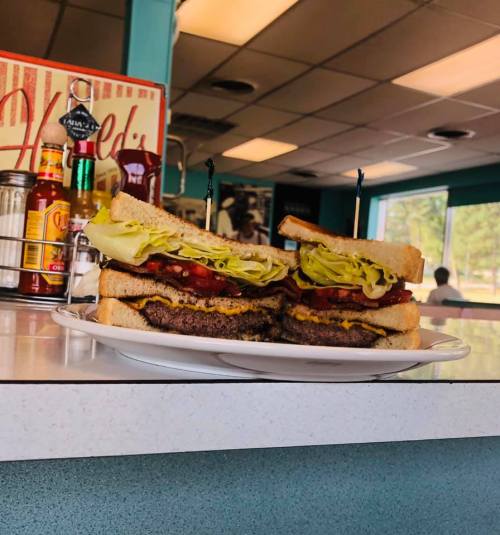 A stacked burger with lettuce, tomato, and cheese on white bread, served on a plate in a diner setting.