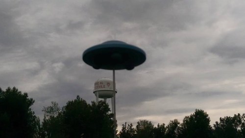 A UFO hovers above a water tower surrounded by trees under a cloudy sky.